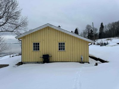 Fotografi av en gul, trepanelert bygning (kanskje uthus eller garasje) sett fra siden. Bygningen står på et snødekt område med vann i bakgrunnen.