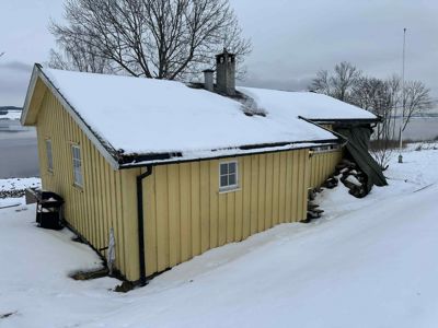 Fotografi av en gul, trepanelert hytte eller liten bygning med snø på taket og i omgivelsene.