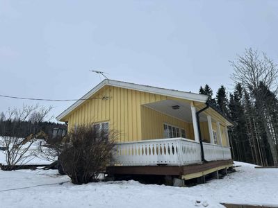 Fotografi av en gul hytte med veranda, sett fra siden, med snø på bakken og skog i bakgrunnen.