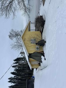 Fotografi av en gul hytte med snø på taket og i omgivelsene, sett fra siden.