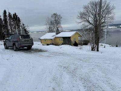 Fotografi av en gul hytte ved vannet i snø, sett fra bakken.