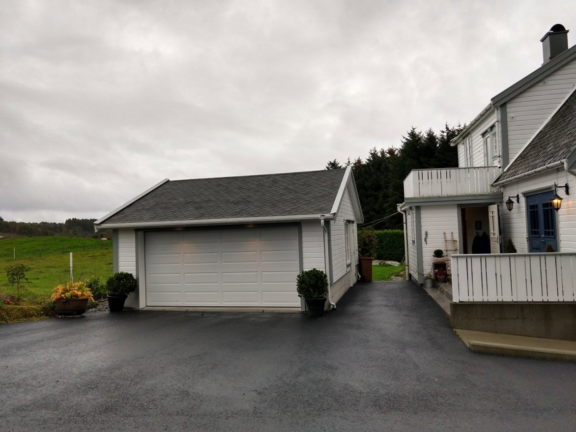 Fotografi av en hvit garasje og en del av en hvit villa sett fra bakkenivå. Bildet viser fasaden, taket, kjøretøysspor og omgivelser.
