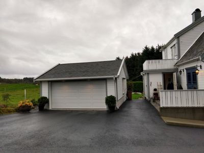 Fotografi av en hvit garasje og en del av en hvit villa sett fra bakkenivå. Bildet viser fasaden, taket, kjøretøysspor og omgivelser.