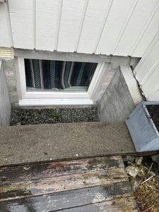 Fotografi av en vindusåpning i en betongfundamentmur, sett fra en trapp nedover. Bildet viser også deler av fasaden, trappetrinn og en planteramme.