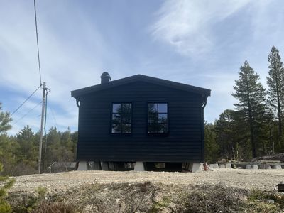 Fotografi av en mørk, ferdig eller nesten ferdig bygget hytte sett fra siden. Bygningen står på betongfundamenter i et naturområde med skog og en elektrisk stolpe.