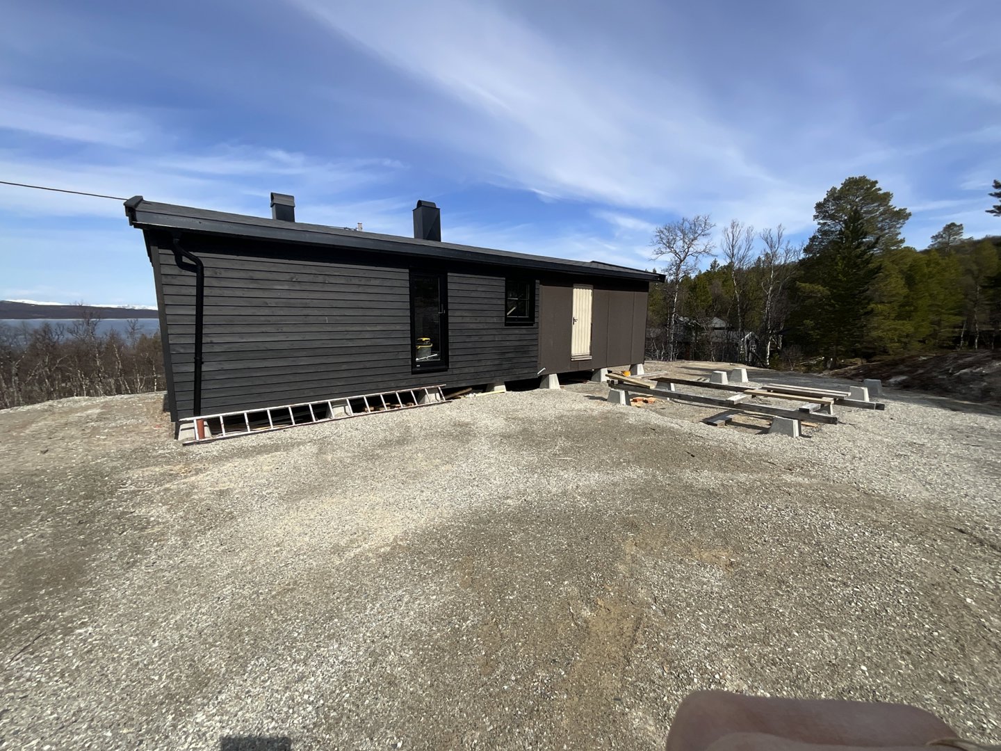 Fotografi av en ferdig eller nesten ferdig bygning (hus) sett fra siden, med utsikt over vann og skog i bakgrunnen.