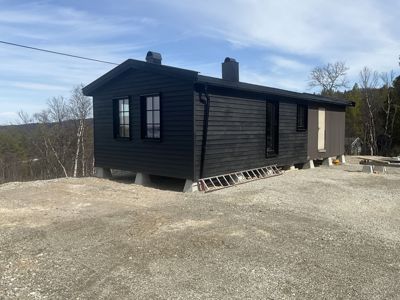 Fotografi av en ferdig eller nesten ferdig bygget hytte sett fra siden. Bygningen har mørke fasader, vinduer og to røykrør, og står på betongfundamenter på en grusete tomt.