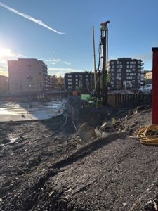 Fotografi av et byggeplass med boremaskin, graving og eksisterende boligblokker i bakgrunnen.