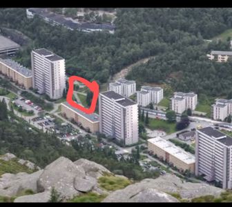 Fotografi tatt fra høyde (f.eks. fra en fjelltopp) som viser et boligområde med flere høyhus og lavere bygninger. Bildet inneholder en håndtegnet rød markering som peker på et spesifikt område mellom to bygninger.