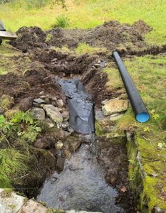 Fotografi av en utgravd grøft på en byggeplass med en svart rørledning og en svart membran (teller) som ligger i grøften.