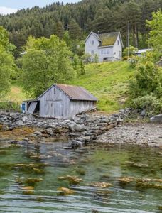 Fotografi av en eksisterende bygning (en gammel uthus) ved vannkanten, med en annen hus i bakgrunnen.