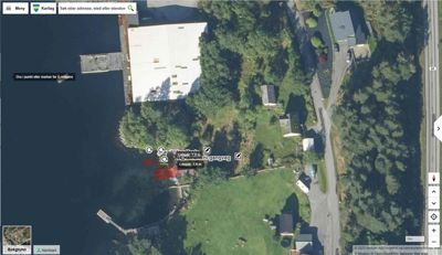 Satellittbilde (kart) fra Google Maps som viser et område med vann, en vei, skog og flere bygninger.