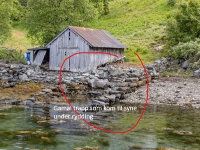 Fotografi av en gammel, grå hytte ved vannet med en steinmur og en markert trapp som ble funnet under rydding.