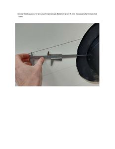 Fotografi av en hånd som holder en målepinsett (caliper) for å måle avstanden til et brennbart materiale på en rørledning.