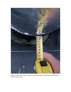 Fotografi som viser en hånd som holder en målebånd for å måle avstanden mellom en pipe og en bunn av blikk-kasse. Bildet illustrerer en fysisk kontroll eller måling på en byggeplass.