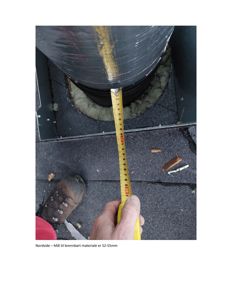 Fotografi av en hånd som holder en målebånd for å måle avstand til brennbart materiale under en røykledning. Bildet viser en fysisk inspeksjon eller kontroll.