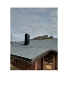 Fotografi av en bygning sett fra siden, som viser taket med en svart røykledning, takkanten, en del av fasaden og bakgrunnen med fjell.