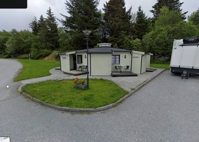 Fotografi av en liten, hvit bygning (muligens en utedo eller hytte) sett fra utsiden, med omgivende natur og asfaltert vei.