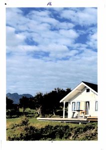 Fotografi av en hvit hytte med veranda, plassert i et grønt landskap med fjell i bakgrunnen.