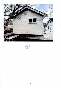 Fotografi av en hvit, liten bygning (kanskje en uthus eller garasje) sett fra siden. Bildet viser fasaden, vinduet, taket og en del av omgivelsene med trær og en annen bygning i bakgrunnen.