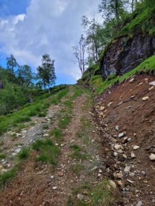 Fotografi av en steinete, grusete sti som går opp en bratt, skogkledt bakke med en steil fjellvegg til høyre.