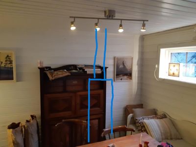 Fotografi av et interiør (spisestue eller stue) med møbler, malerier og en lysarmatur. Det er tegnet blå linjer på bildet som indikerer en vegg eller romdeling.