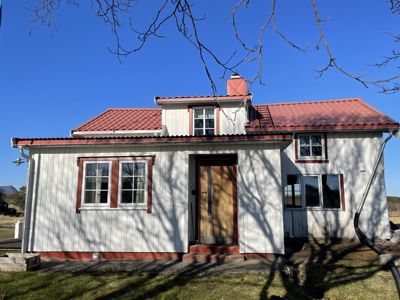 Fotografi av en hvit, trepanelt enebolig med rød takflate og røde vindusrammer, sett fra front.