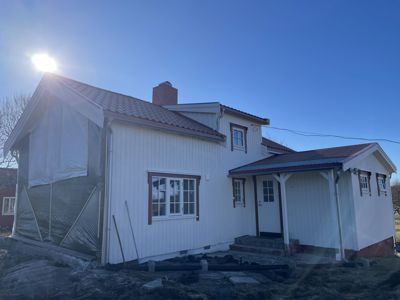 Fotografi av en hvit villa under pågående utvendig renovering eller vedlikehold, med solskin og blå himmel.