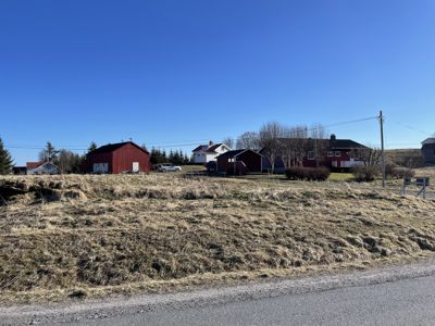 Fotografi av en eksisterende bygning eller tomt sett fra en vei, med rød skur og hus i bakgrunnen.