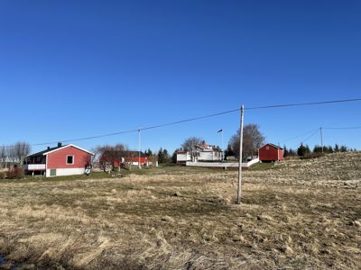 Fotografi av et boligfelt med flere røde hus og en åker i forgrunnen under blå himmel.