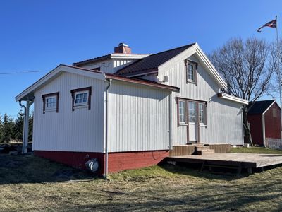Fotografi av en hvit, panelt husfasade med rød grunnmur, sett fra siden. Bildet viser også en del av tomme, trær og en annen rød bygning i bakgrunnen.