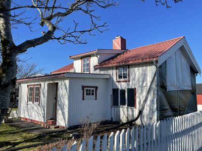 Fotografi av en hvit, trepanelert enebolig med rød takflate. Bygningen er delvis dekket av beskyttelsesplast, noe som antyder at det pågår eller har pågått renovering. Bildet viser fasaden og en del av tommeområdet med en hvit hekk.