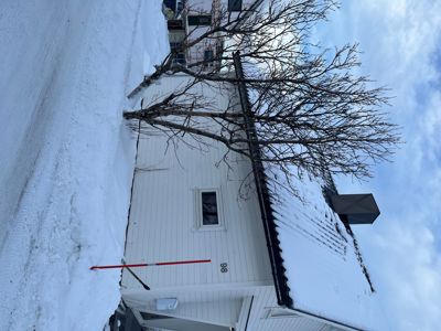 Fotografi av en hvit, enetasjes bolig med snø på taket og bakken. Bildet viser fasaden, en trær, en rød markørstang og deler av nabolaget.