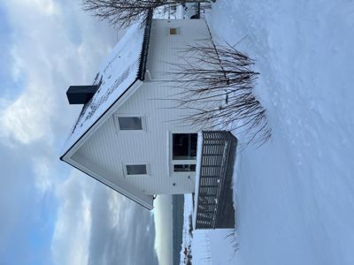 Fotografi av en hvit, trepanelert hytte med snø på taket og i omgivelsene, sett fra siden.