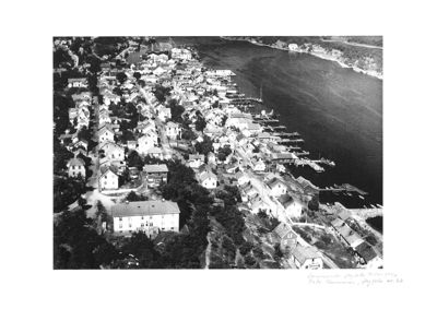 Et svart-hvitt flyfoto som viser et tettsted langs en kystlinje med mange hus, båter og en større bygning i forgrunnen.