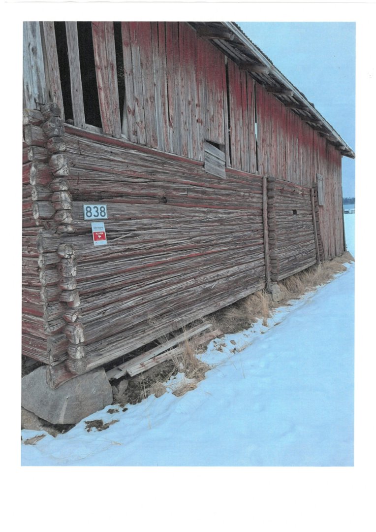 Fotografi av en gammel, rød og grå tømmerbygning (sannsynligvis et uthus eller skur) sett fra siden. Bygningen står på steinfundament i snø.
