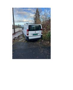 Fotografi av en hvit varebil parkert på en grus- eller asfaltert overflate ved en vei eller eiendom.