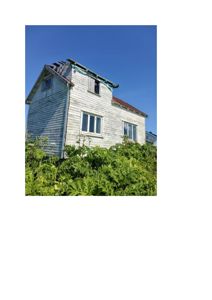 Fotografi av en gammel, hvitmalt trehytte sett fra siden og litt underfra. Bygningen har et skrått tak, vinduer og er omgitt av grønt. Bildet viser en eksisterende bygning.