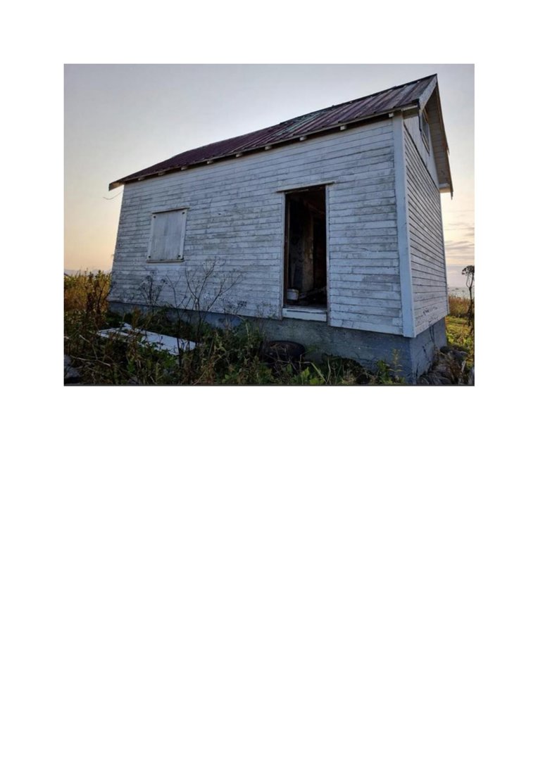 Fotografi av en liten, hvit, utvunnet hytte eller uthus med et rustfarget tak, sett fra en vinkel som viser både front og sidevegg.