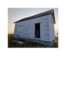 Fotografi av en liten, hvit, utvunnet hytte eller uthus med et rustfarget tak, sett fra en vinkel som viser både front og sidevegg.