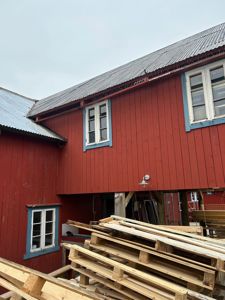 Fotografi av en rød, trepanelt bygning med blå vindusrammer og et bølget tak. Bildet viser fasaden og en del av konstruksjonen under bygningens utstikkende del.