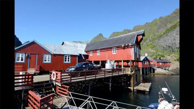 Fotografi av røde stuer på stoler ved kysten, med bil og båter i forgrunnen.