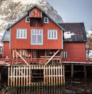 Fotografi av en rød, tradisjonell bygning (kanskje en hytte eller fiskeribod) på stolper ved vannet, med fjell i bakgrunnen.
