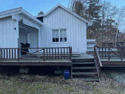 Fotografi av en hvit, trepanelert hytte sett fra bakkenivå. Bildet viser bygningens fasade, en stor terrasse med trapper, og omgivelsene med skog i bakgrunnen.