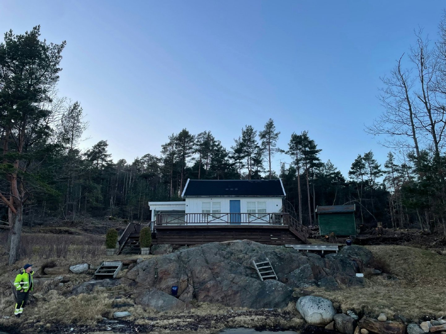 Fotografi av en hvit hytte med mørk tak og terrasse, plassert på en steinete bakke i skog. En person i sikkerhetsvest står i forgrunnen.