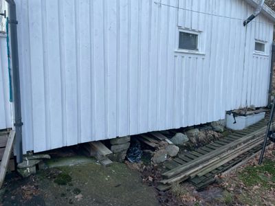 Fotografi av en hvit, paneltett bygning sett fra siden, som viser fasaden, vinduer, nedløp og fundamentet med steiner og planker.