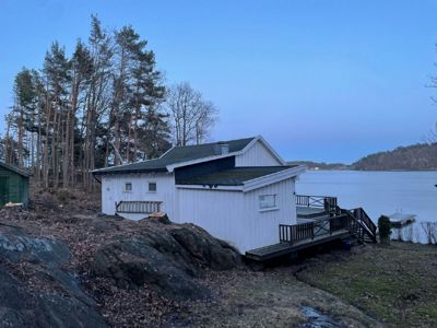 Fotografi av en hvit hytte ved vannet med en utsikt over en skog og en fjord.