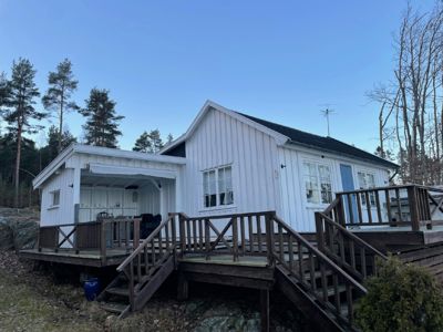 Fotografi av en hvit, trepanelt hytte med en stor terrasse og trapper, sett fra bakken mot en blå himmel.