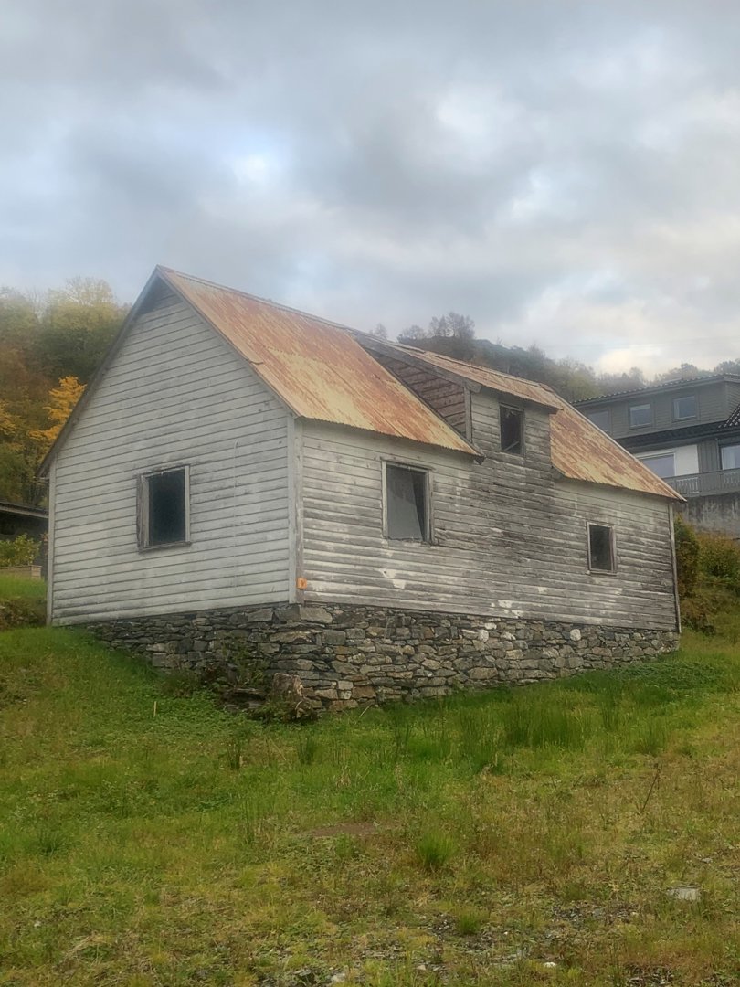Fotografi av en gammel, hvitmalt trebygning med rustet tak og steinmur, sett fra en skrå vinkel.