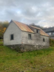 Fotografi av en gammel, hvitmalt trebygning med rustet tak og steinmur, sett fra en skrå vinkel.